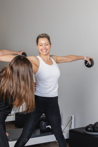 Woman raising weighted balls to the side while a trainer checks her posture
