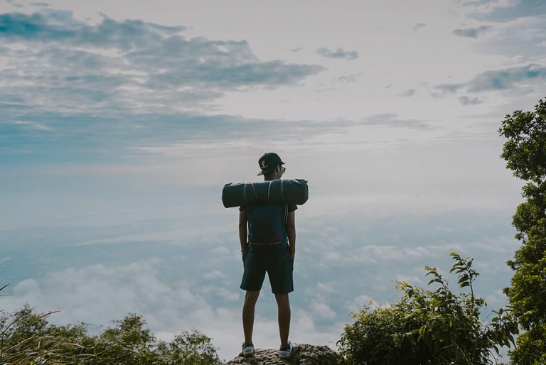 Hiker with a rolled mat on their pack standing on a rocky outcrop above the clouds